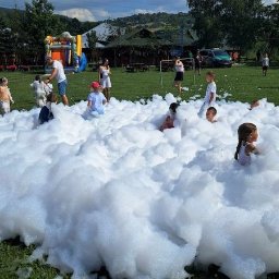 Plenerowo.com Atrakcje & Gastronomia - Dzieci bawią się w pianie na trawie, w tle dmuchany zamek i budynki. Letnia impreza plenerowa z animacjami dla dzieci.