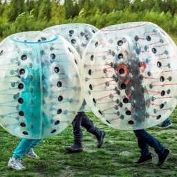 Plenerowo.com Atrakcje & Gastronomia - Trzy osoby w przezroczystych kulach Bumper Ball na zielonej trawie. Widoczne nogi i buty uczestników zabawy. W tle drzewa i krzewy. Dynamiczna scena aktywności na świeżym powietrzu.