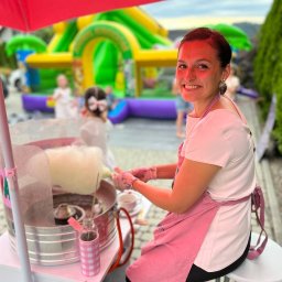 Plenerowo.com Atrakcje & Gastronomia - Uśmiechnięta kobieta w różowym fartuchu robi watę cukrową na wózku, w tle dmuchany zamek i dzieci na imprezie plenerowej. Jasne, naturalne światło.