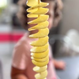 Plenerowo.com Atrakcje & Gastronomia - Spiralny ziemniak na patyku, lekko rozmyte tło z postacią dziecka w różowej koszulce. Jasne, naturalne światło. Nietypowy przysmak na imprezę.
