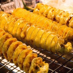 Plenerowo.com Atrakcje & Gastronomia - Spiralne ziemniaki na patyku, ułożone na ruszcie. Widoczne przyprawy w słoikach w tle. Ciepłe oświetlenie podkreśla złocisty kolor przekąski.