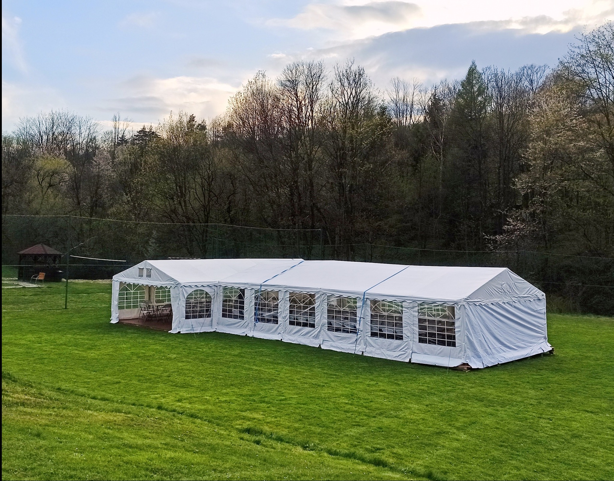 Rozłożony biały namiot imprezowy z oknami na zielonej trawie, w tle drzewa i altana. Widok z góry, lekko z boku. Namiot z wyposażeniem w środku.