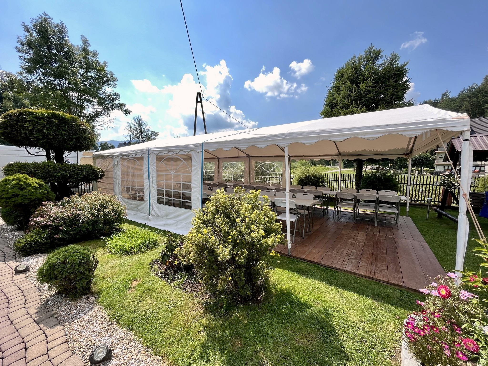 Biały namiot imprezowy z drewnianą podłogą i krzesłami, otoczony zielenią ogrodu w słoneczny dzień. Widoczne krzewy i kwiaty.