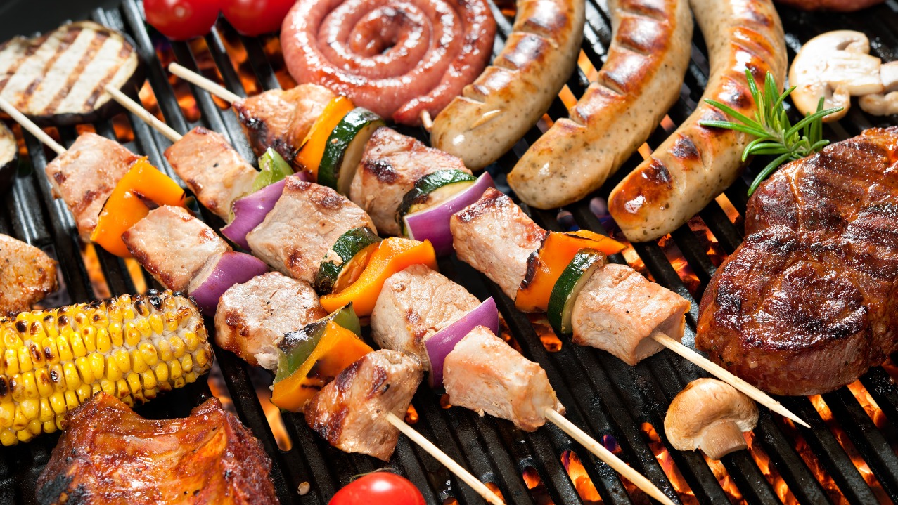 Grillowany stek, kiełbaski i szaszłyki z warzywami na ruszcie. Kukurydza i żeberka w tle. Widoczne płomienie ognia. Idealne na imprezę plenerową.