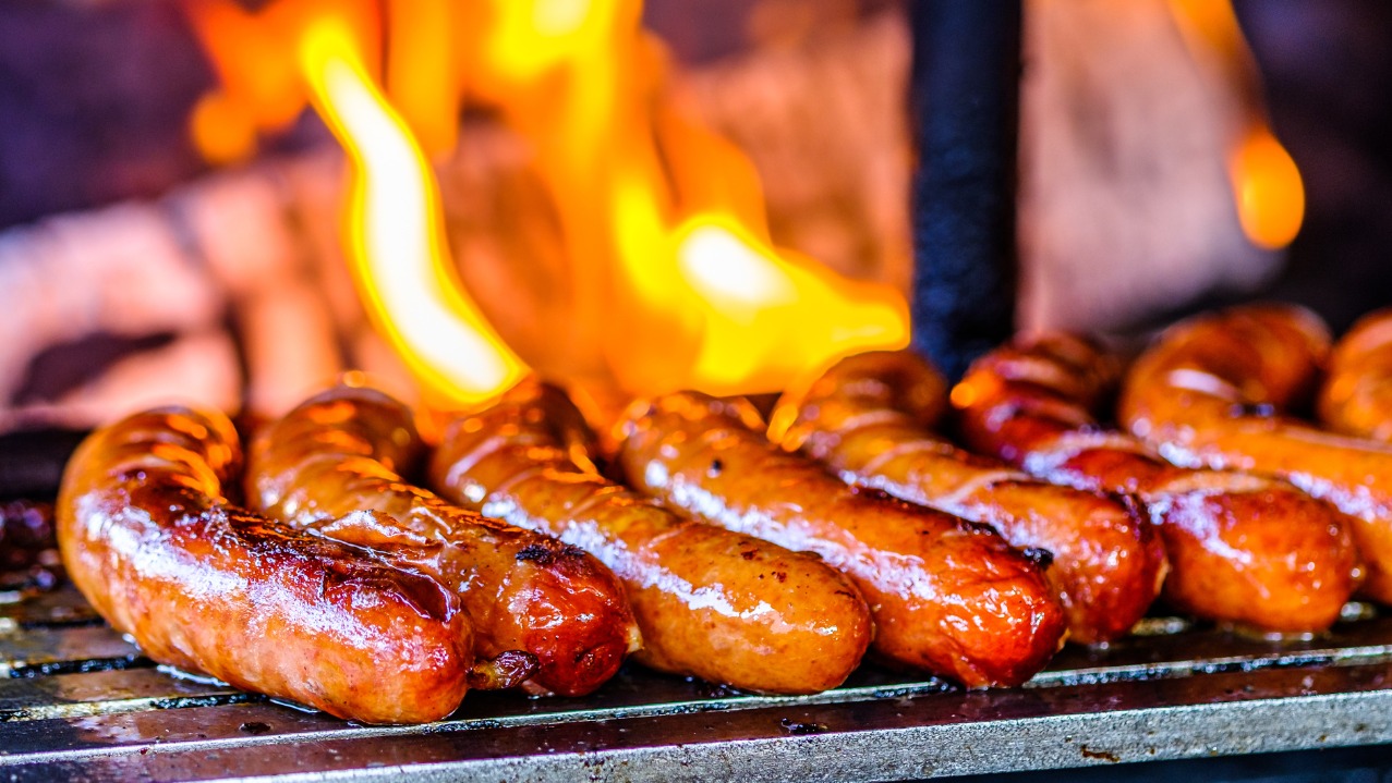 Soczyste kiełbaski na grillu, płomienie tańczą nad nimi, tworząc apetyczny widok. Idealne na imprezę plenerową z wypożyczonym sprzętem.