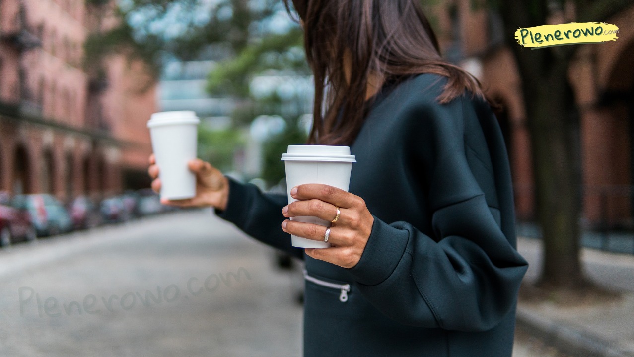 Kobieta na tle miejskiej ulicy trzyma dwa białe kubki z kawą. Ubrana w ciemną bluzę, na palcach widoczne pierścionki. Rozmyte budynki i drzewa w tle.