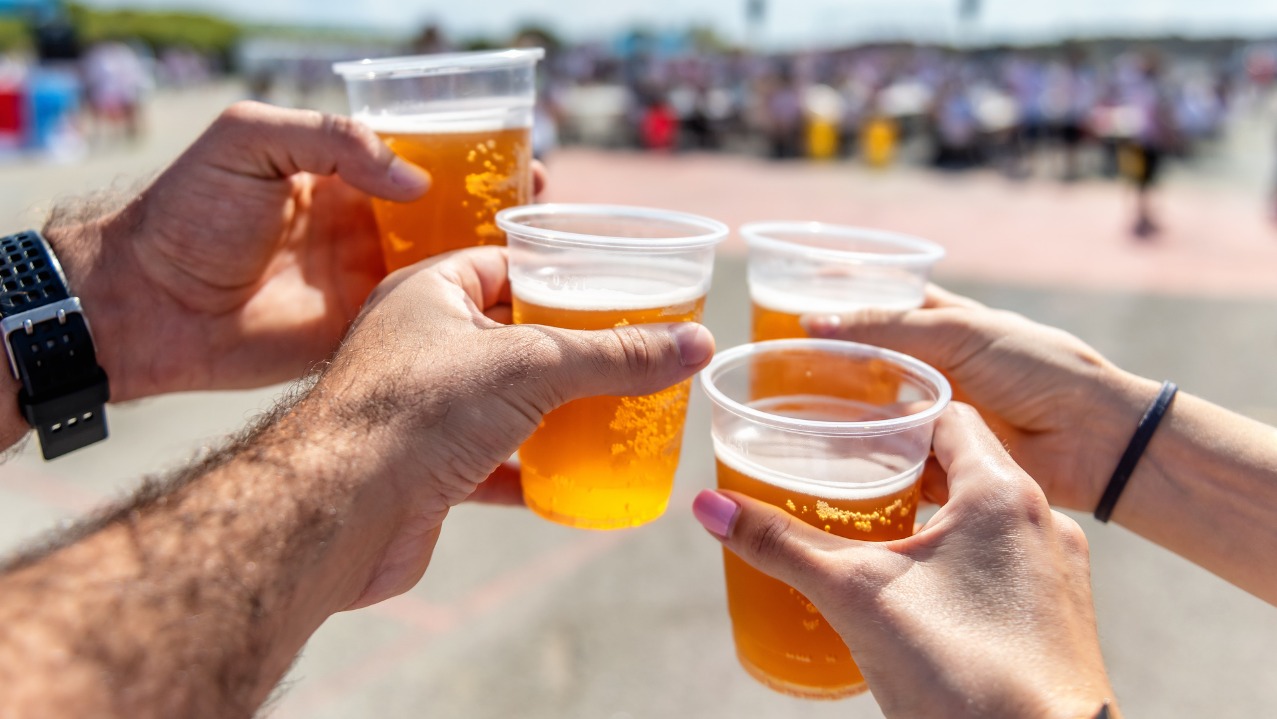 Cztery osoby wznoszą toast piwem w plastikowych kubkach na tle rozmytego tłumu, prawdopodobnie podczas imprezy plenerowej. Widoczne dłonie z bliska.