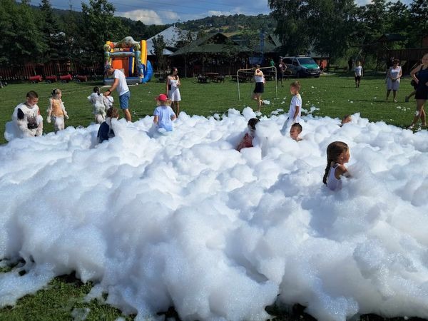 Dzieci bawią się w pianie na trawie, w tle dmuchany zamek i budynki. Letnia impreza plenerowa z animacjami dla dzieci.