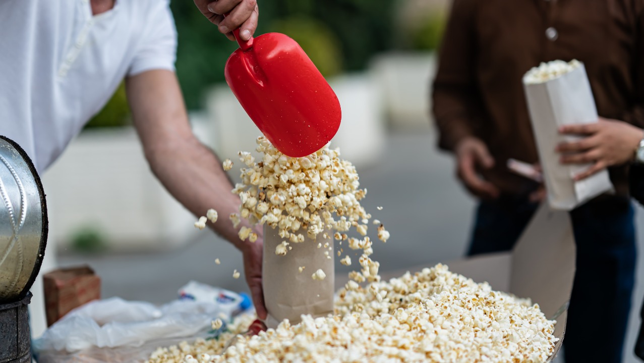 Napełnianie papierowej torby popcornem za pomocą czerwonej łopatki. W tle osoba z torbą popcornu. Widoczny wózek z popcornem i zapas.