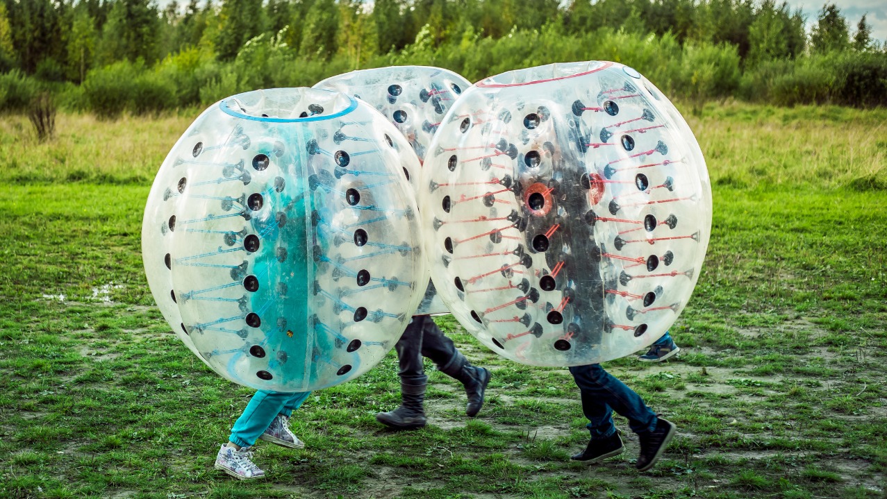 Trzy osoby w przezroczystych kulach Bumper Ball na zielonej trawie. Widoczne nogi i buty uczestników zabawy. W tle drzewa i krzewy. Dynamiczna scena aktywności na świeżym powietrzu.