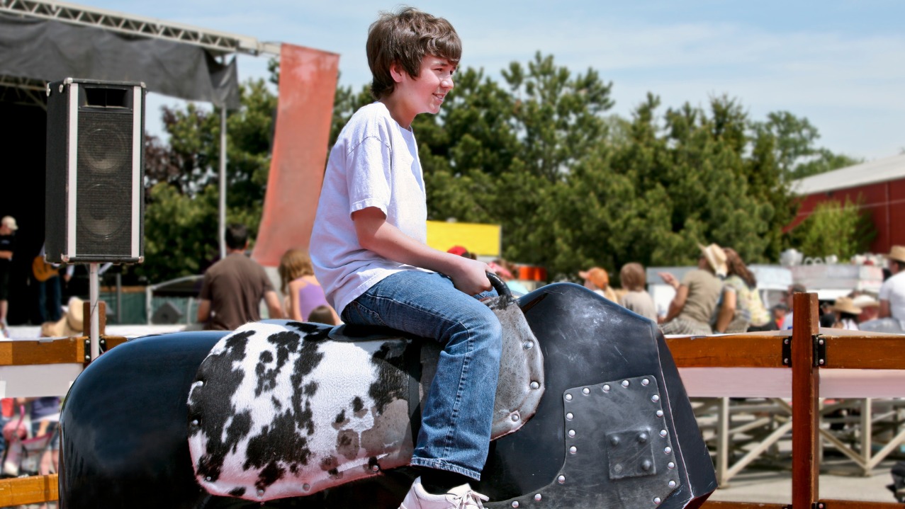 Chłopiec na mechanicznym byku na tle sceny i publiczności. Atrakcja eventowa z imitacją zwierzęcia, zapewniająca rozrywkę podczas imprezy plenerowej.