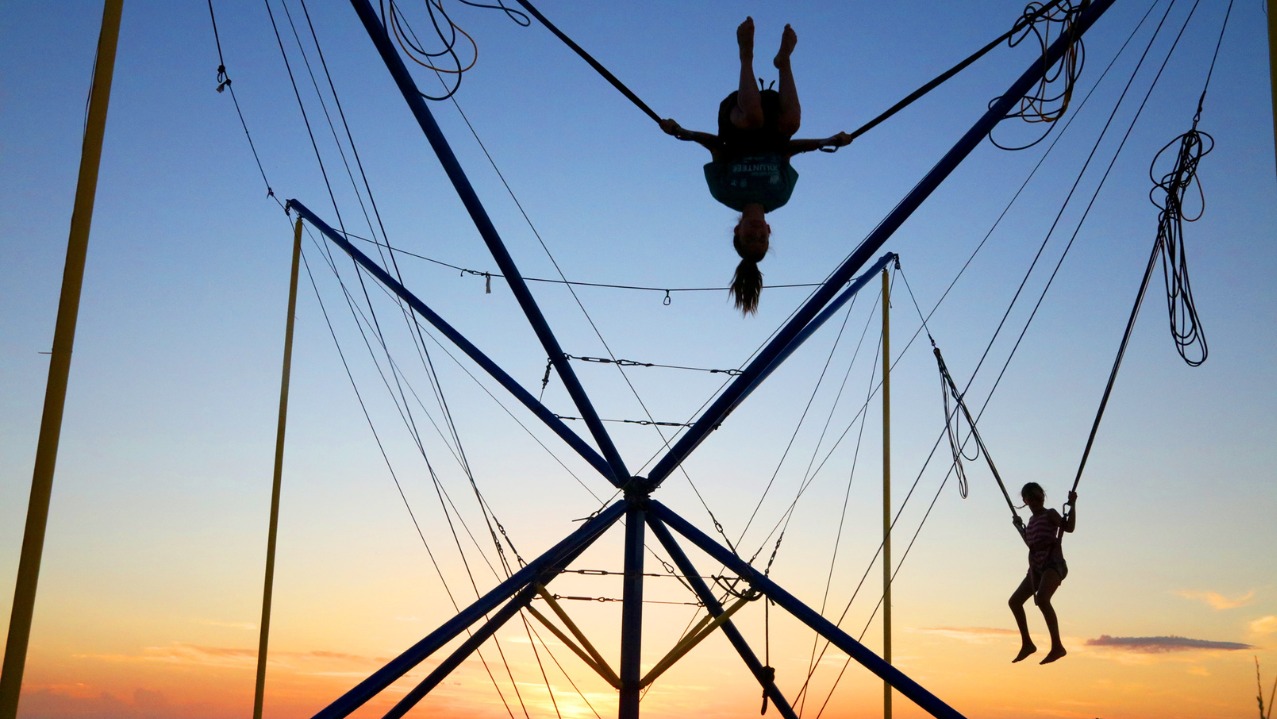 Dwie osoby na bungee trampolinie na tle zachodzącego słońca. Jedna osoba w powietrzu, druga skacze. Konstrukcja z lin i metalowych rurek.