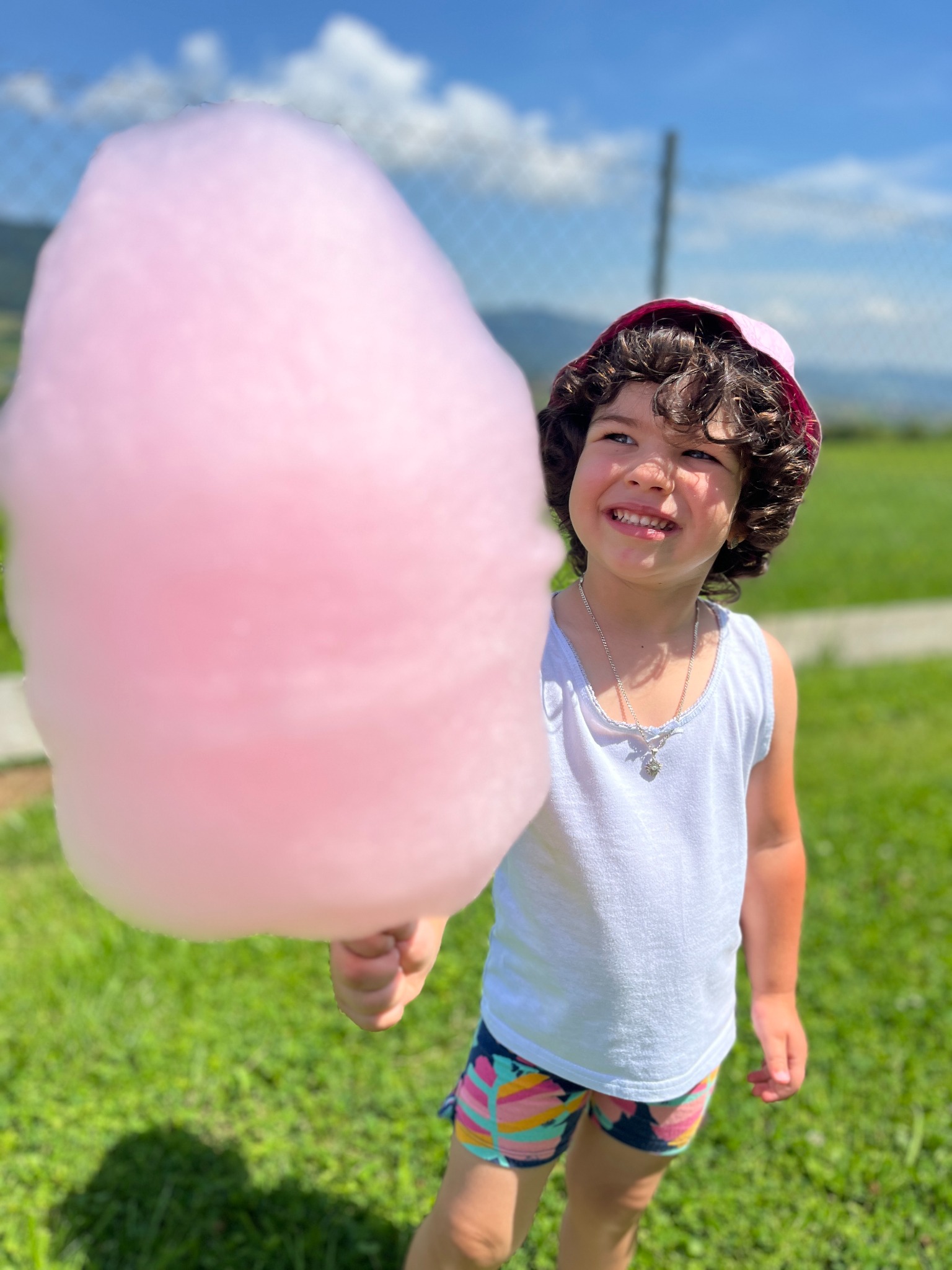 Uśmiechnięta dziewczynka w różowym kapeluszu trzyma watę cukrową na tle zielonej trawy i błękitnego nieba z chmurami.