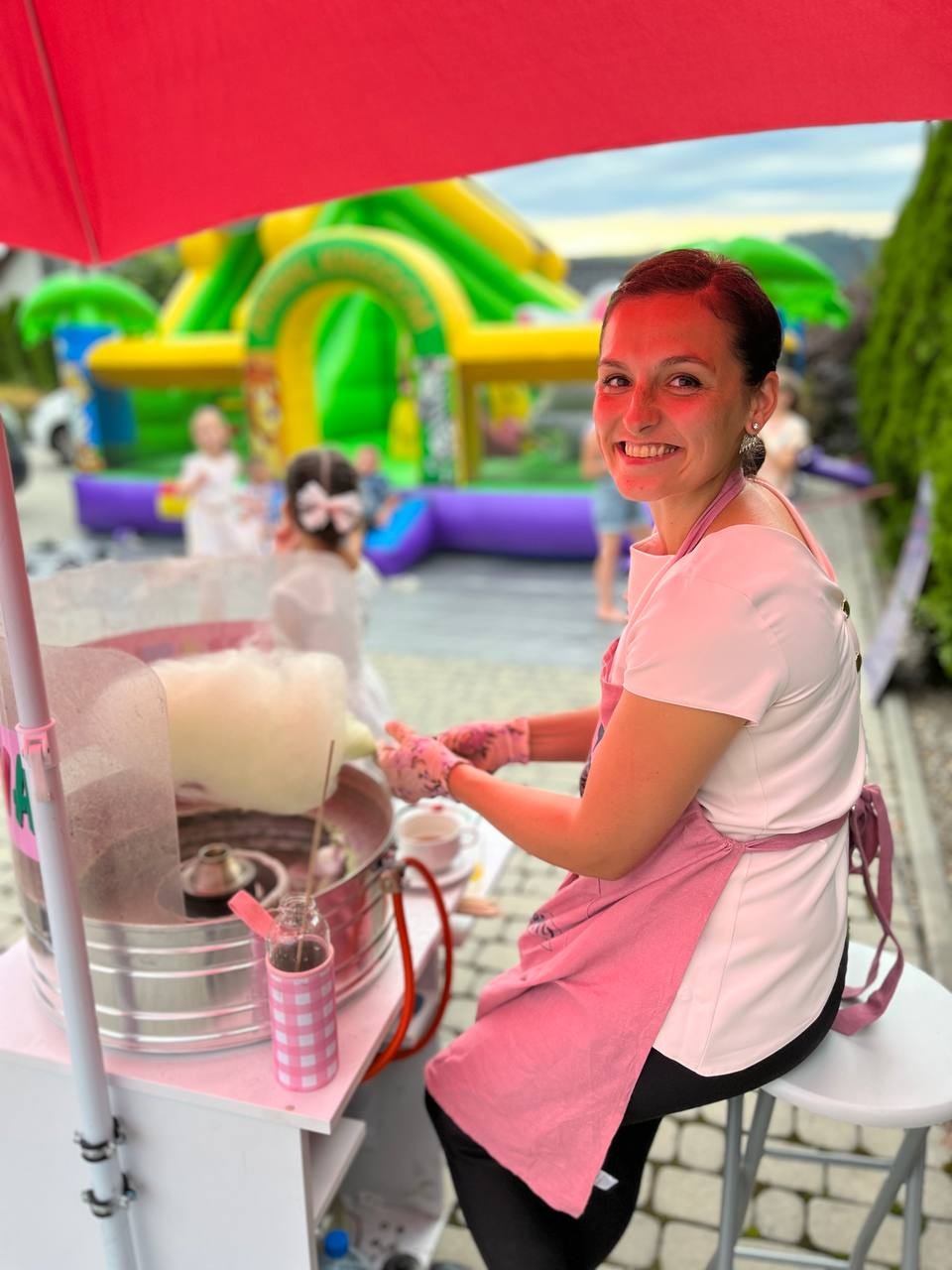 Uśmiechnięta kobieta w różowym fartuchu robi watę cukrową na wózku, w tle dmuchany zamek i dzieci na imprezie plenerowej. Jasne, naturalne światło.