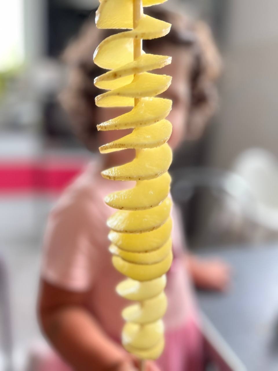 Spiralny ziemniak na patyku, lekko rozmyte tło z postacią dziecka w różowej koszulce. Jasne, naturalne światło. Nietypowy przysmak na imprezę.