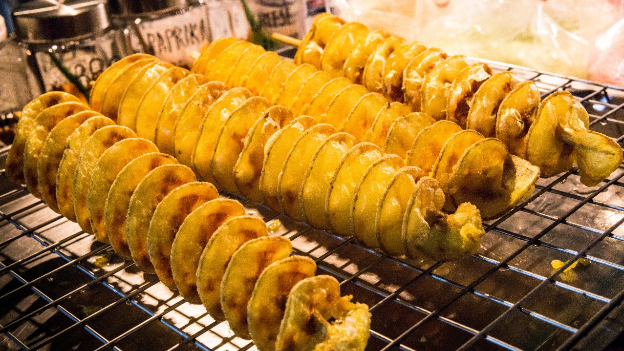 Spiralne ziemniaki na patyku, ułożone na ruszcie. Widoczne przyprawy w słoikach w tle. Ciepłe oświetlenie podkreśla złocisty kolor przekąski.