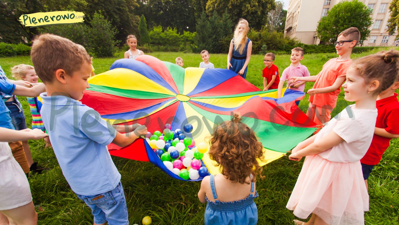 Grupa dzieci bawi się na trawie, trzymając kolorową chustę animacyjną wypełnioną piłeczkami. W tle widoczna kobieta i budynek.
