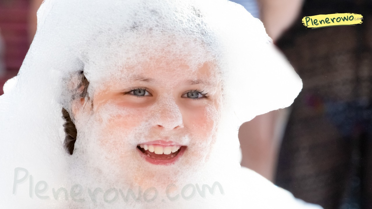 Uśmiechnięta dziewczynka z twarzą i włosami pokrytymi pianą podczas imprezy plenerowej z użyciem wytwornicy piany.