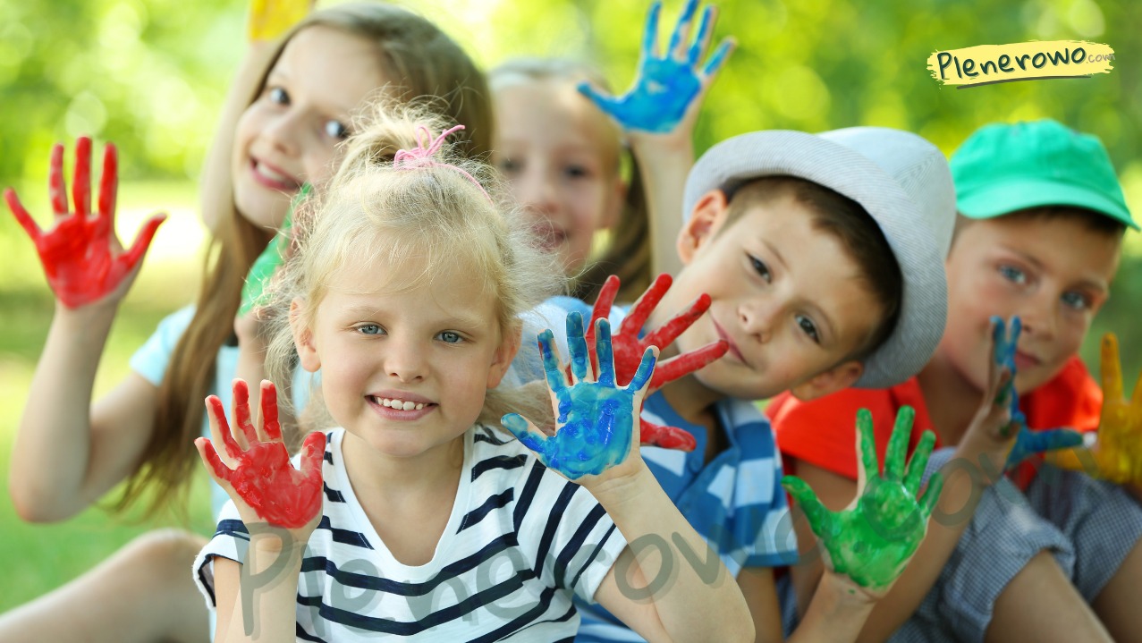 Uśmiechnięte dzieci z pomalowanymi dłońmi w kolorach czerwonym, niebieskim i zielonym, siedzące na trawie. Plenerowe warsztaty artystyczne.