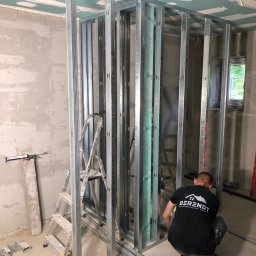 Berendt wykończenia wnętrz i remonty - Montaż metalowej konstrukcji ścianki działowej z płyt karton-gips. Widoczna drabina, narzędzia i pracownik w koszulce z logo firmy wykończeniowej.