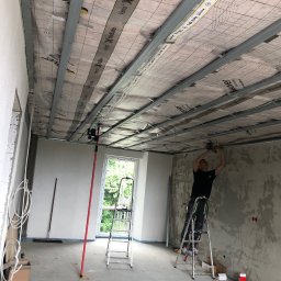 Berendt wykończenia wnętrz i remonty - Montaż sufitu podwieszanego: pracownik na drabinie mocuje profile do izolacji. Widoczny laser krzyżowy, narzędzia i materiały budowlane w surowym wnętrzu.