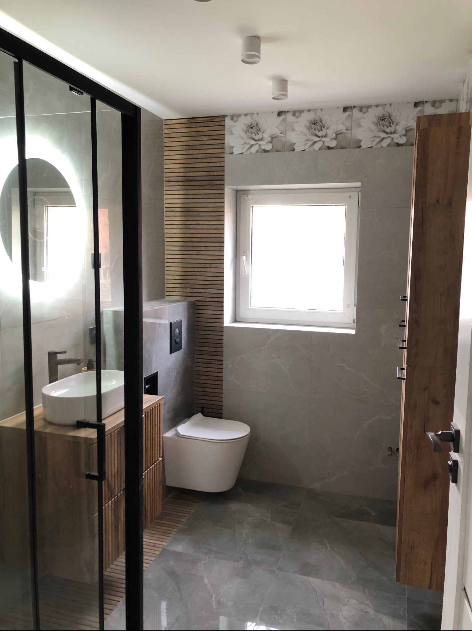 Nowoczesna łazienka z kabiną prysznicową typu walk-in, umywalką nablatową na drewnianej szafce i toaletą w zabudowie. Szare płytki imitujące marmur i drewniane akcenty.