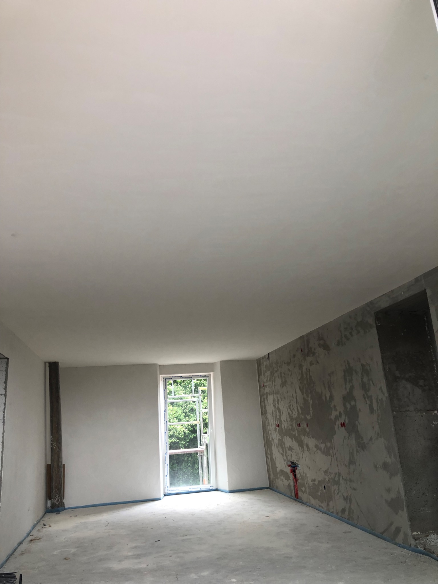 Surowe wnętrze w trakcie prac wykończeniowych: gładzie na ścianach, okno z widokiem na zieleń, przygotowane podłoże. Widoczne ślady po instalacjach.