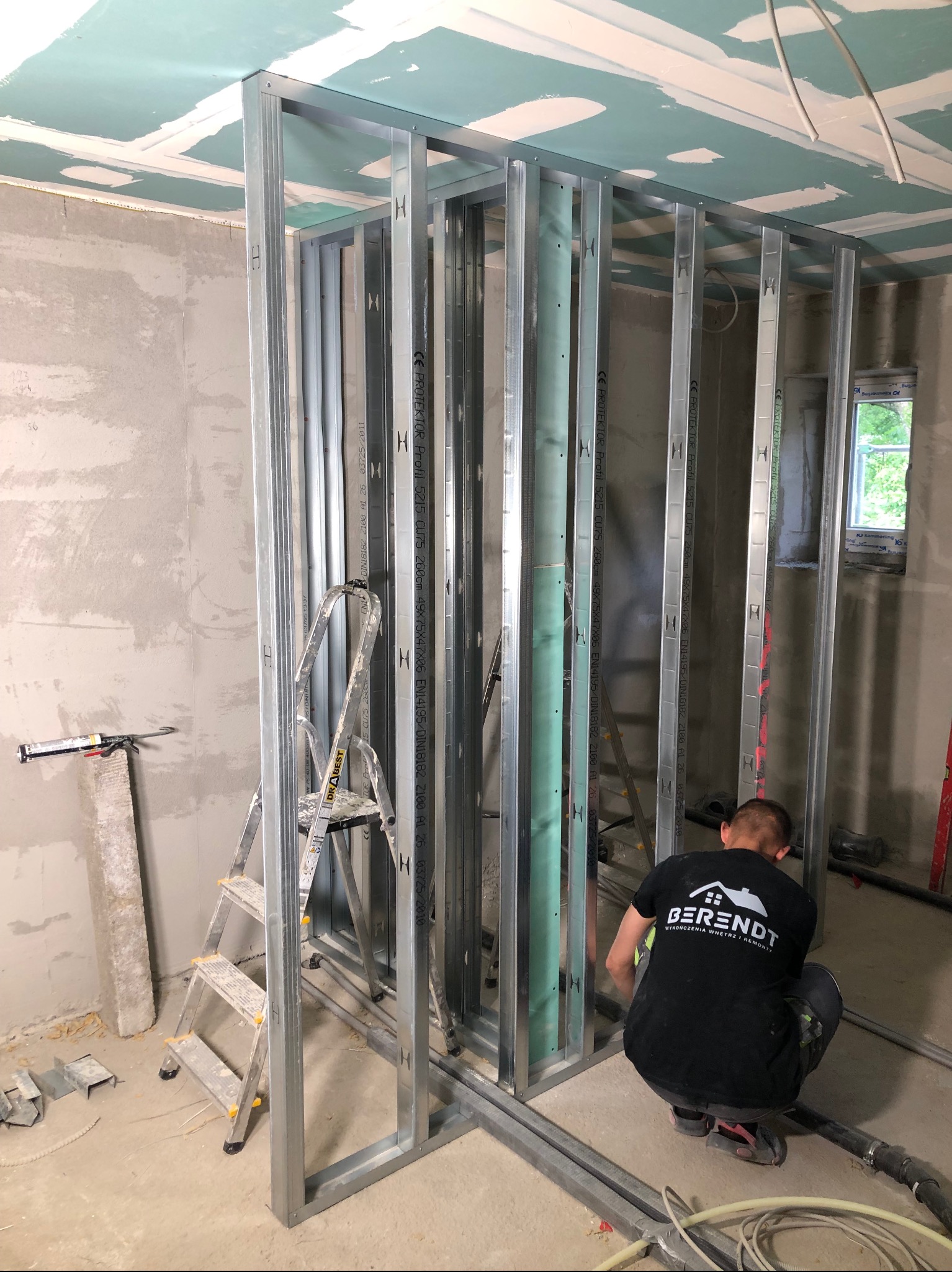 Montaż metalowej konstrukcji ścianki działowej z płyt karton-gips. Widoczna drabina, narzędzia i pracownik w koszulce z logo firmy wykończeniowej.