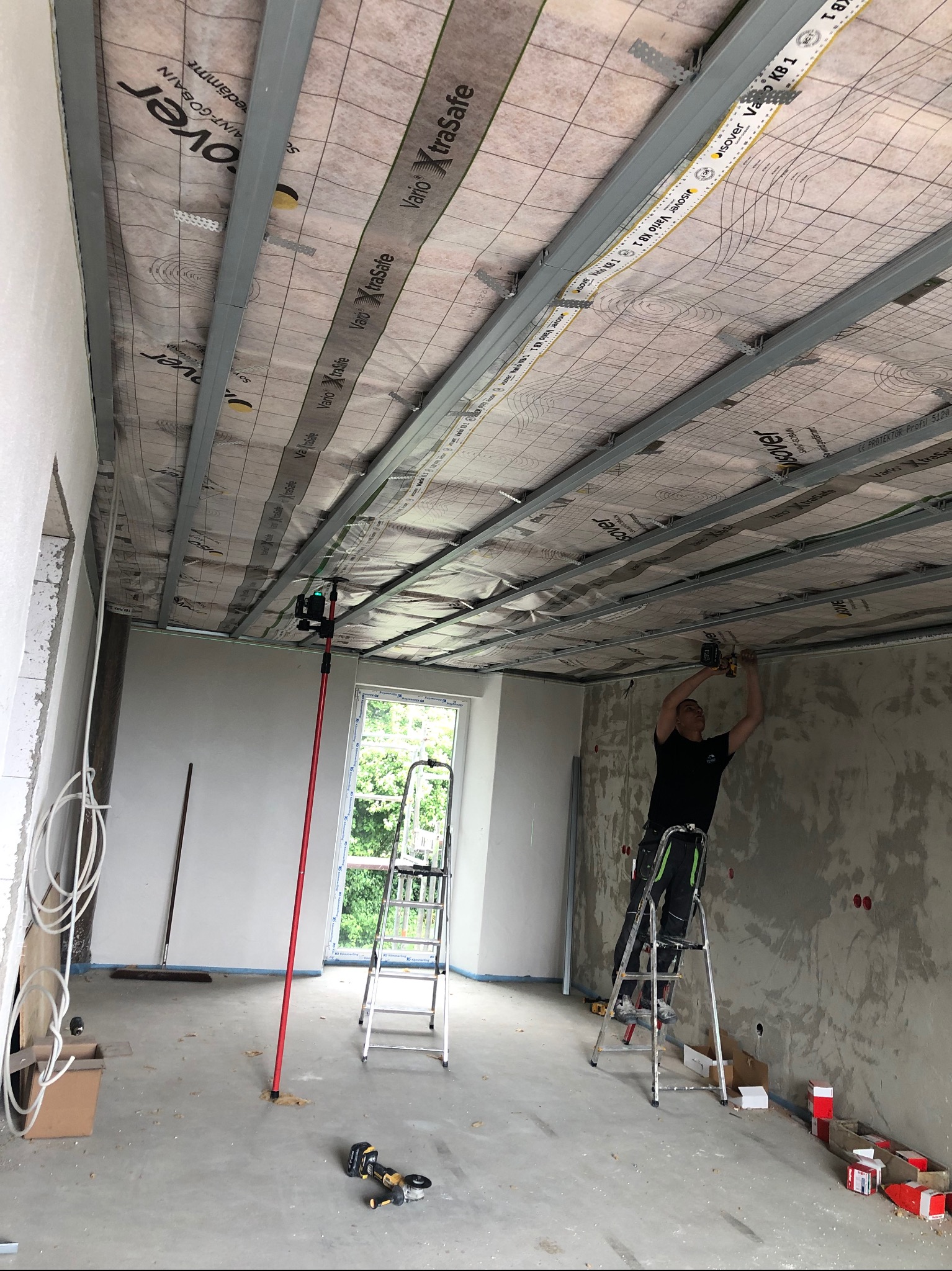Montaż sufitu podwieszanego: pracownik na drabinie mocuje profile do izolacji. Widoczny laser krzyżowy, narzędzia i materiały budowlane w surowym wnętrzu.
