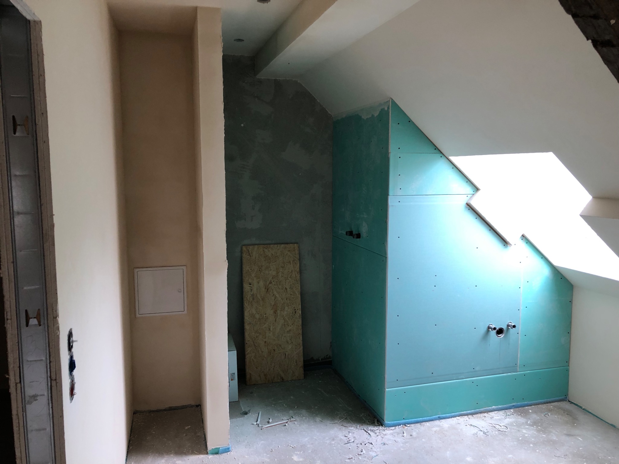 Poddasze w trakcie adaptacji: ściany z surowym tynkiem, zabudowa z płyty karton-gips w kolorze turkusowym, widoczne rury instalacji wodnej, przygotowane do wykończenia.