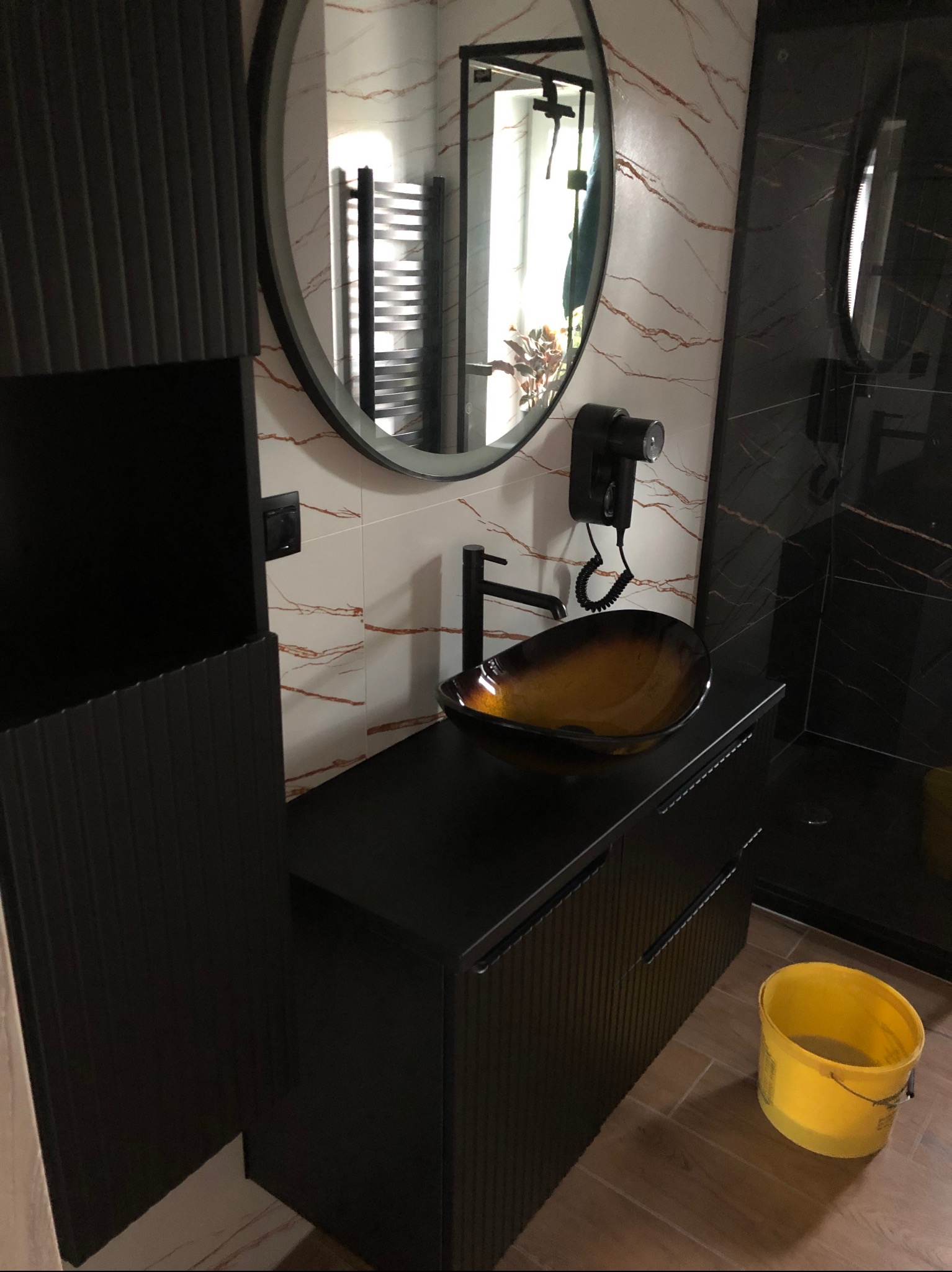 Nowoczesna łazienka z czarną szafką umywalkową, okrągłym lustrem i dekoracyjną umywalką. Widoczny fragment kabiny prysznicowej i wiadro.