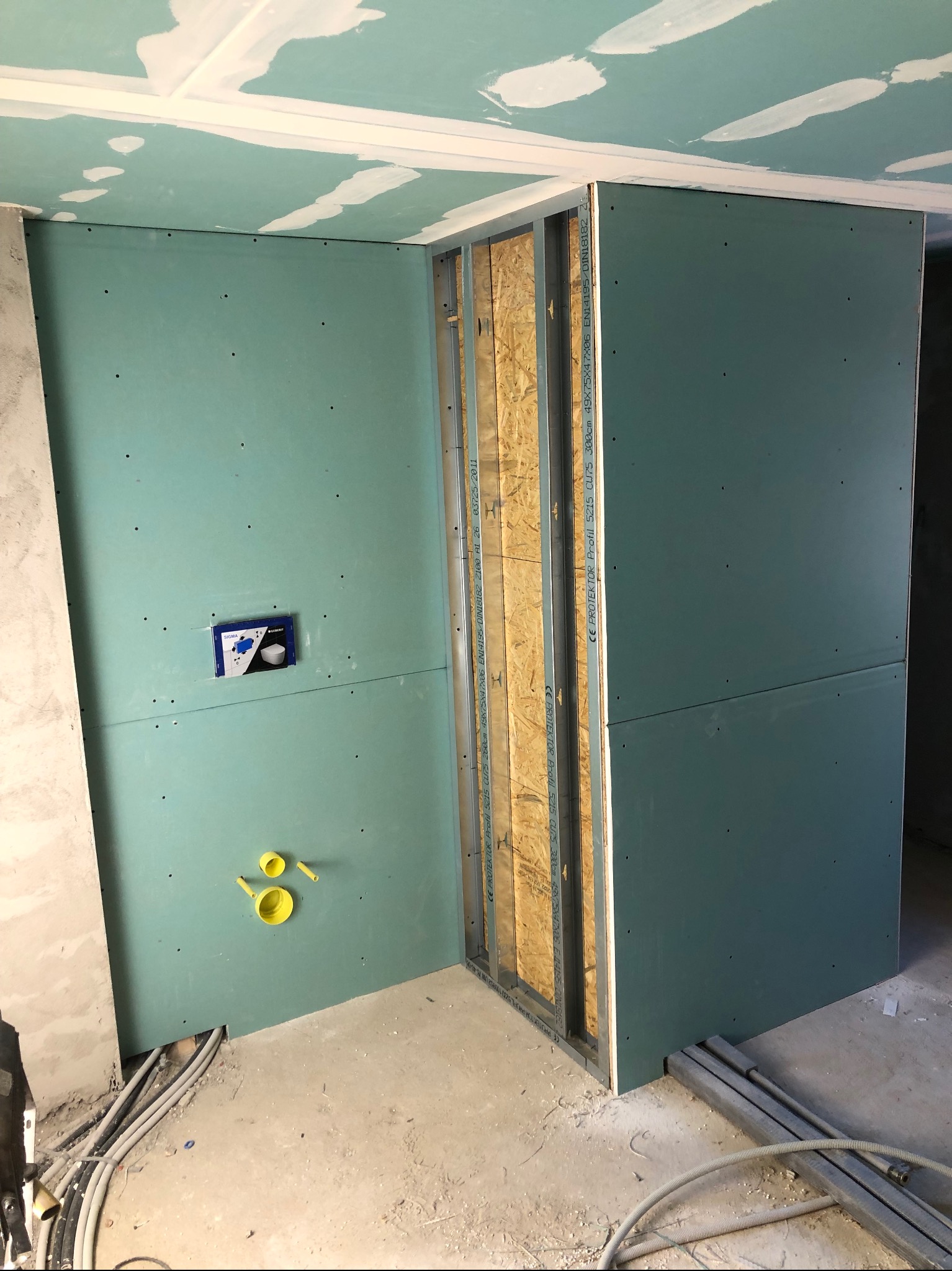 Ściana z płyty karton-gips w trakcie montażu, widoczne profile metalowe i instalacja wodno-kanalizacyjna. Surowy stan, przygotowanie do wykończenia.