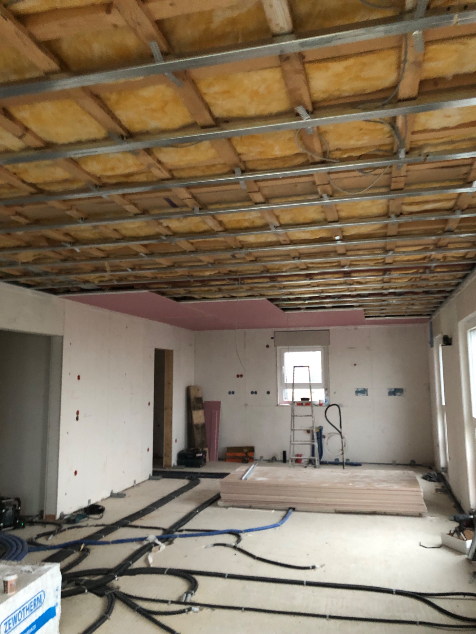 Wnętrze w trakcie montażu sufitu podwieszanego z izolacją. Widoczne profile, wełna mineralna, częściowo zamontowane płyty gipsowe i instalacje na podłodze.