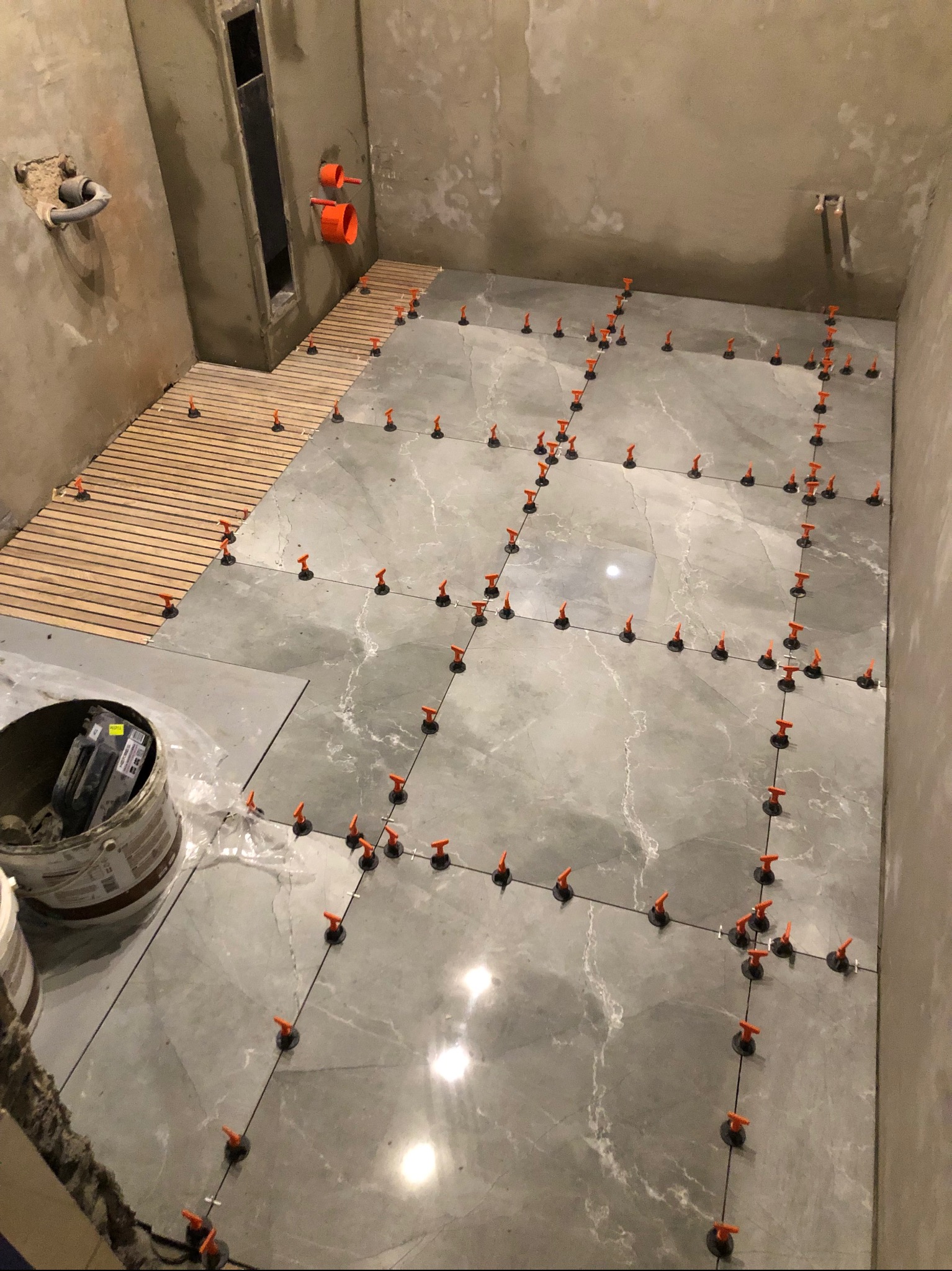 Układanie szarych płytek podłogowych z marmurkowym wzorem w łazience, z użyciem pomarańczowych klipsów poziomujących. Częściowo ułożona drewniana podłoga w kabinie prysznicowej.