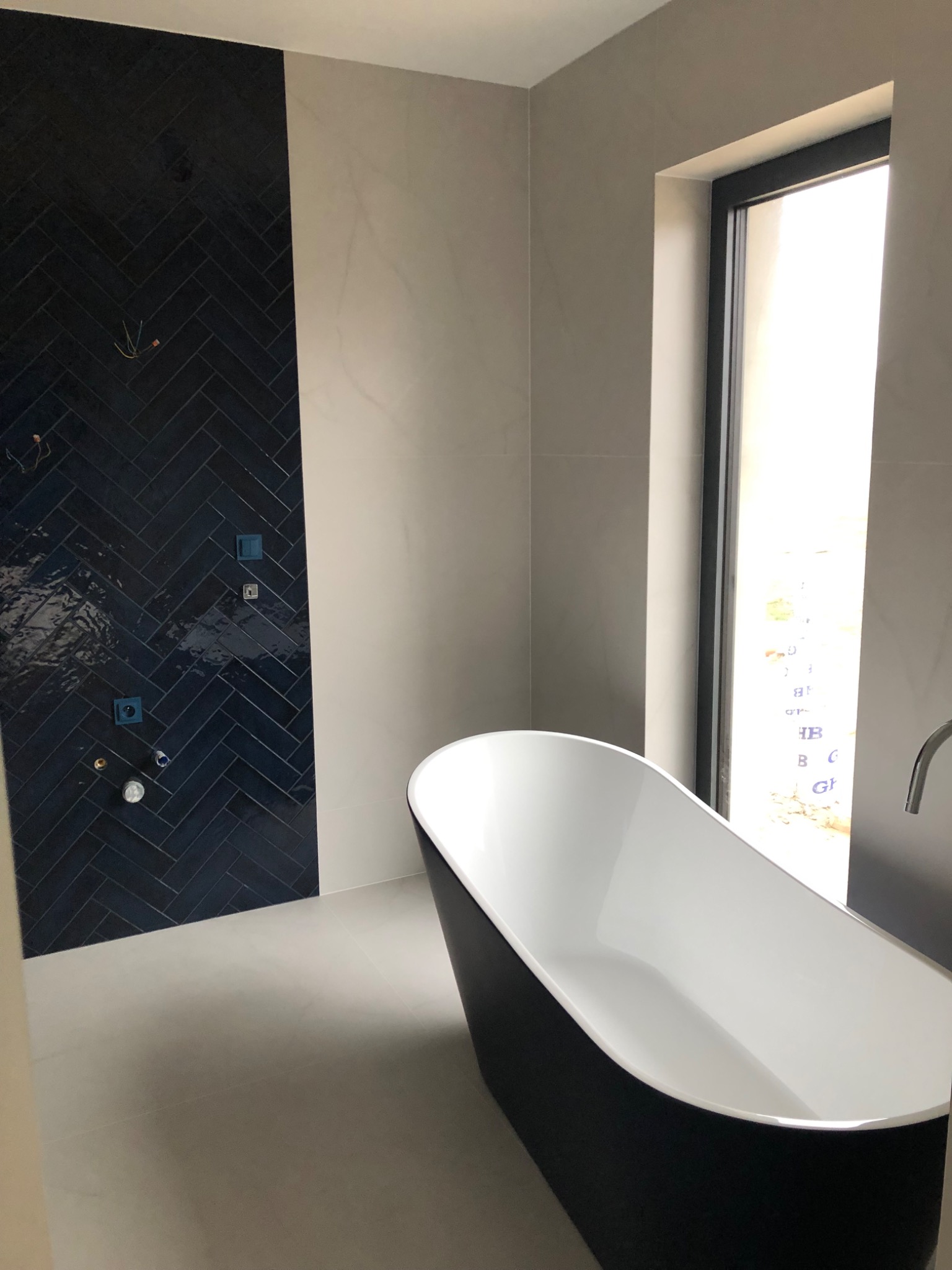 Nowoczesna łazienka z czarną wanną wolnostojącą i ścianą wyłożoną ciemnymi, błyszczącymi płytkami ułożonymi w jodełkę. Minimalistyczny design, jasne płytki podłogowe.