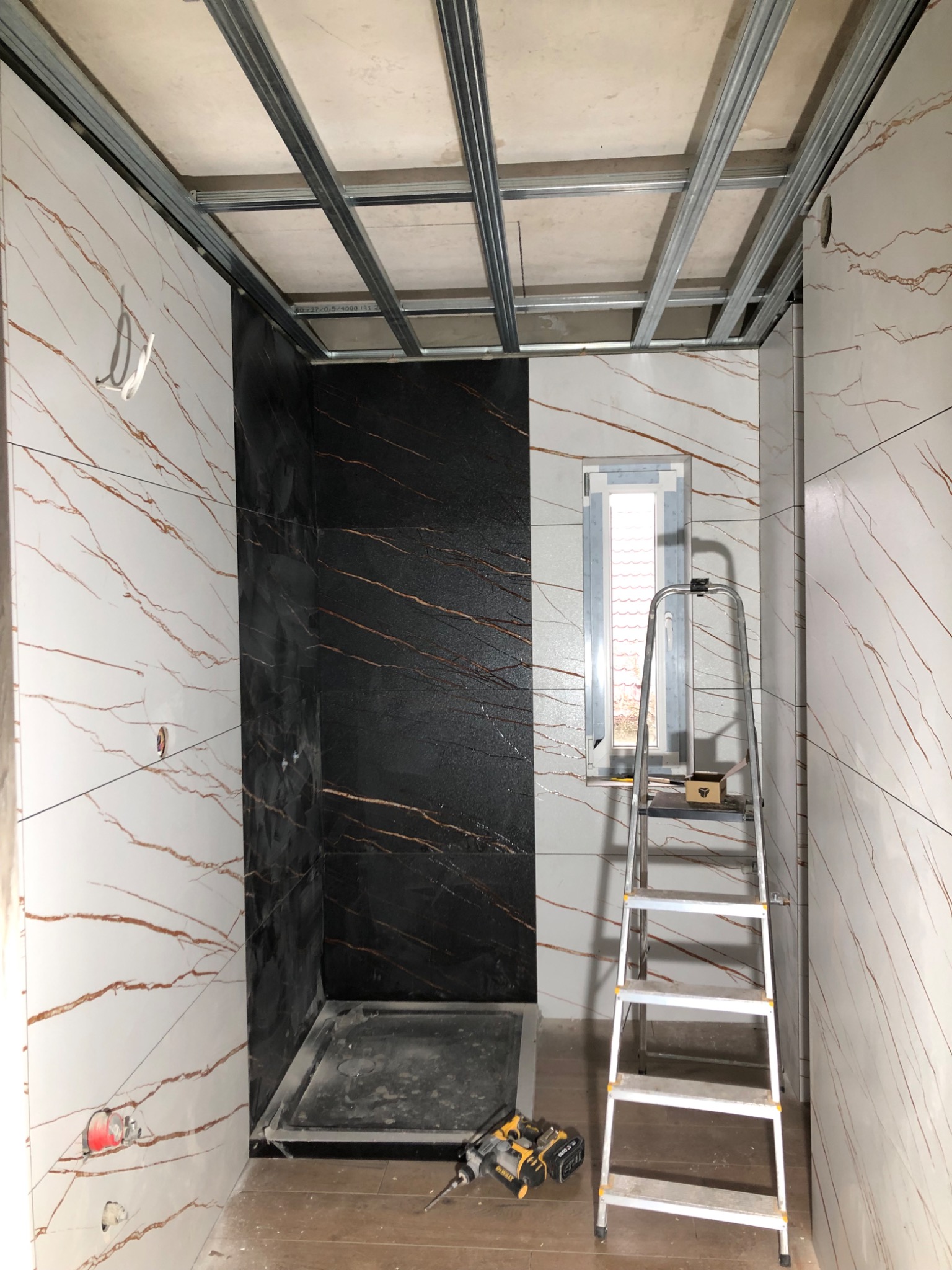 Łazienka w trakcie wykańczania: białe i czarne płytki imitujące marmur, metalowa konstrukcja sufitu podwieszanego, drabina i narzędzia na podłodze.