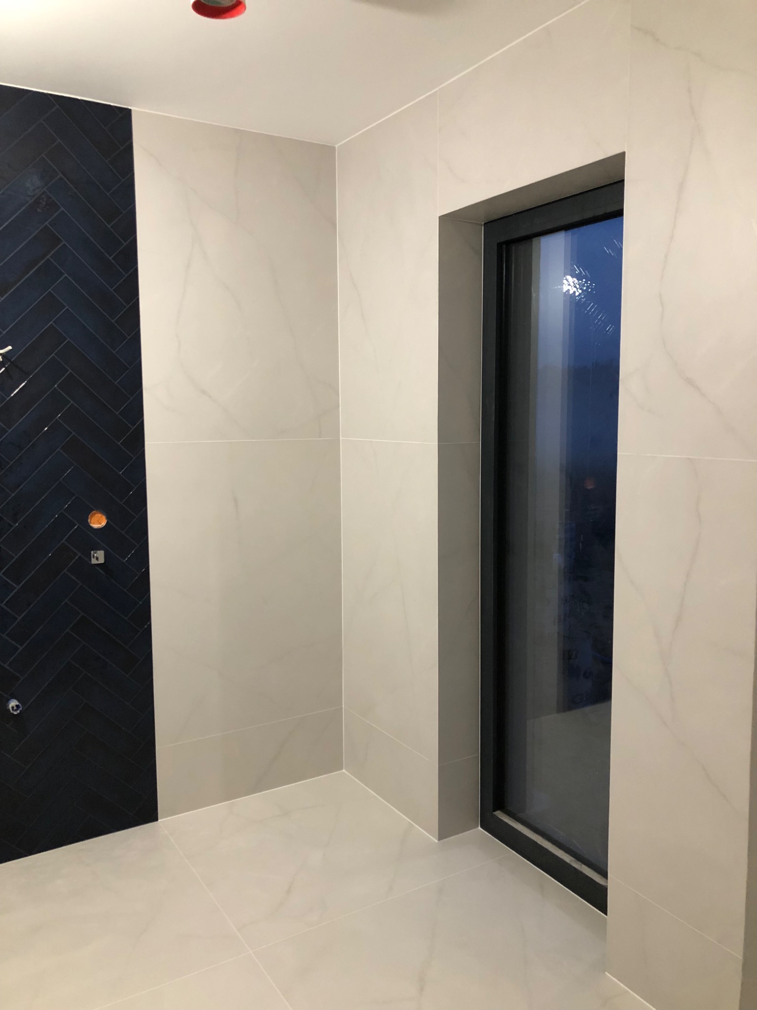 Wykończona łazienka z płytkami w odcieniach bieli i granatu ułożonymi w jodełkę, widoczne okno z czarną ramą i fragment instalacji.