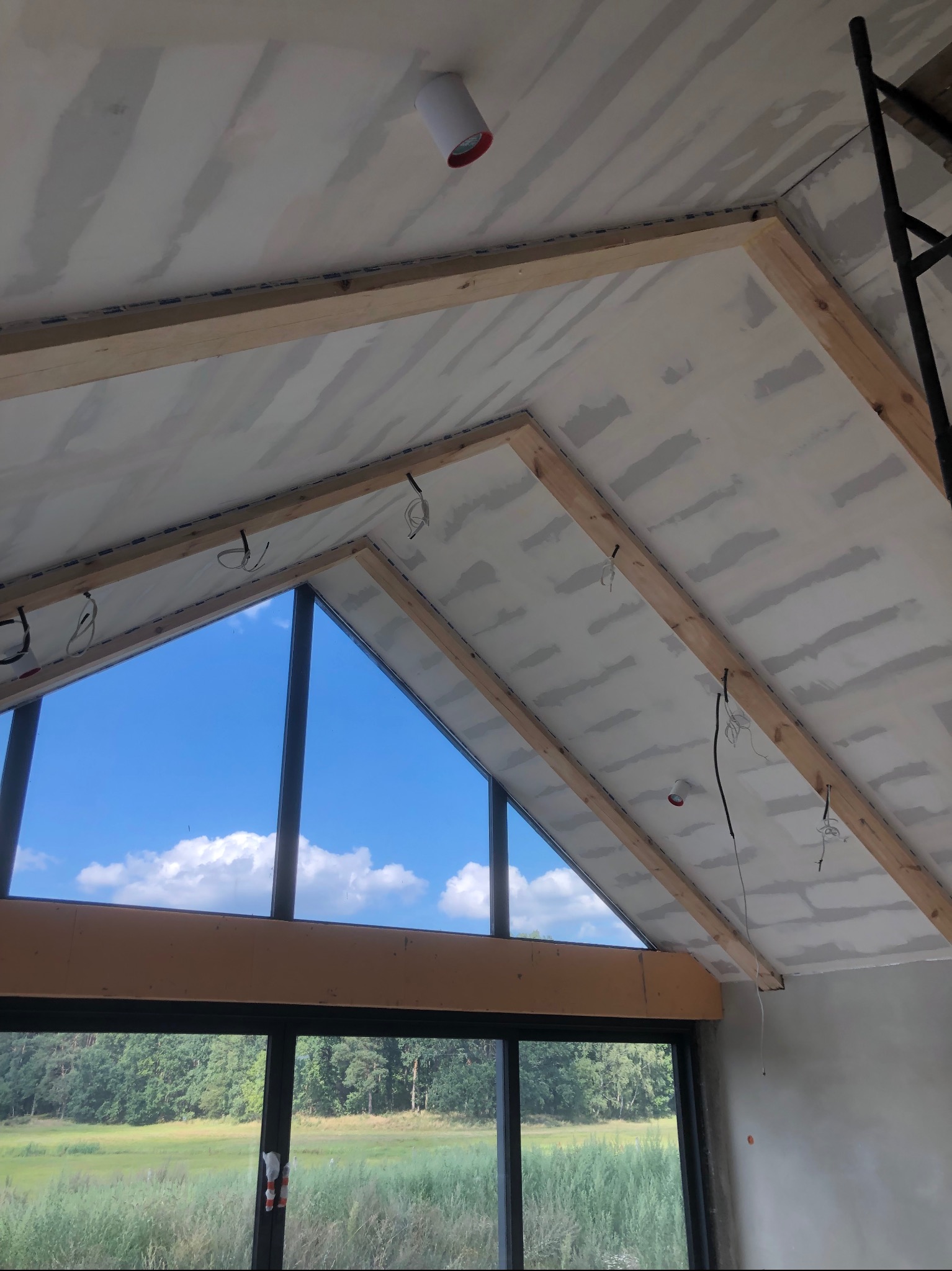 Skośny sufit z płyt kartonowo-gipsowych, drewniane belki konstrukcyjne, widoczne przewody elektryczne, okno z widokiem na zieleń, etap prac wykończeniowych.