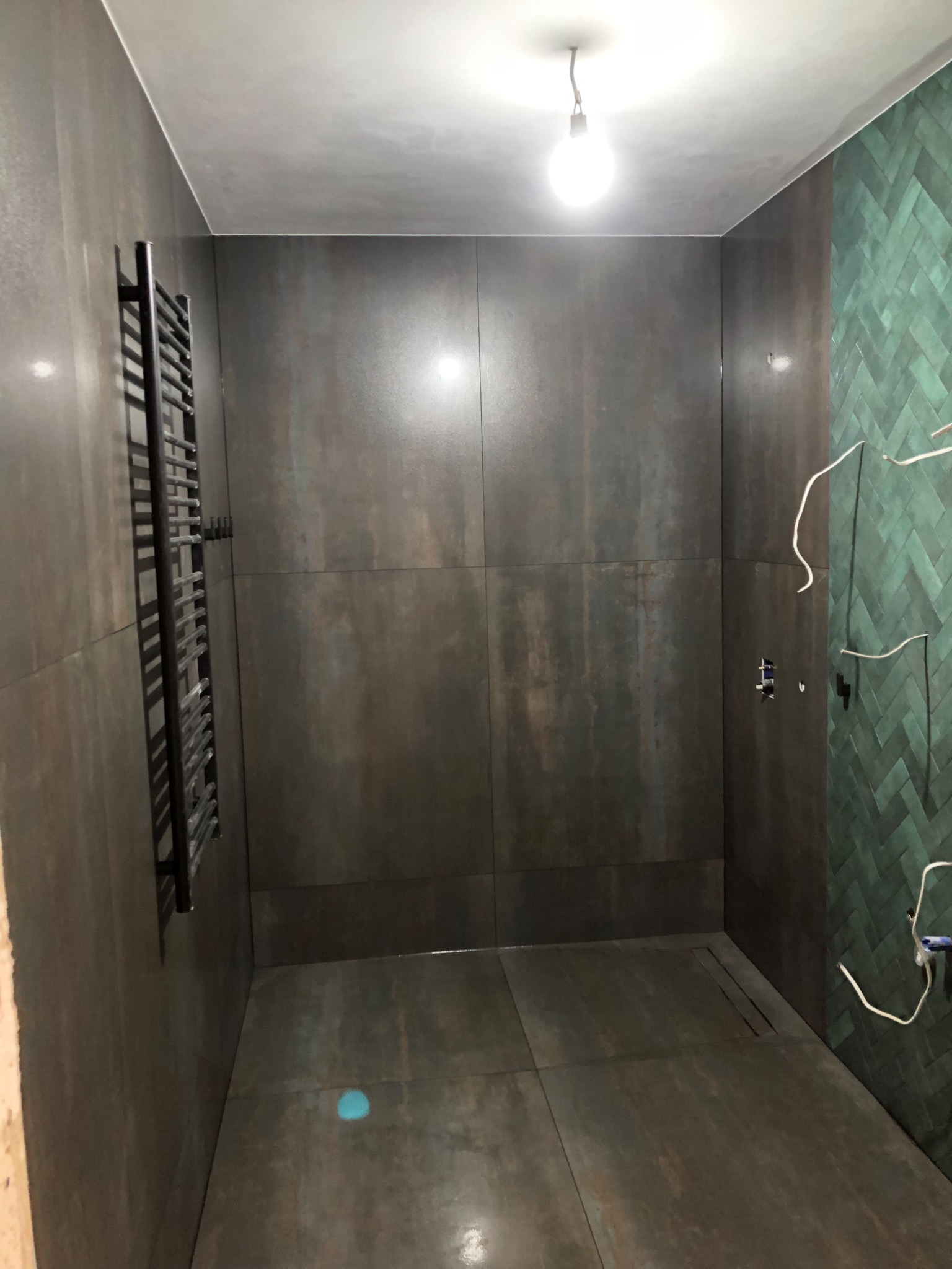Nowoczesna łazienka z ciemnymi płytkami imitującymi metal, zielona ściana w jodełkę i grzejnik drabinkowy. Widoczne niedokończone instalacje elektryczne.