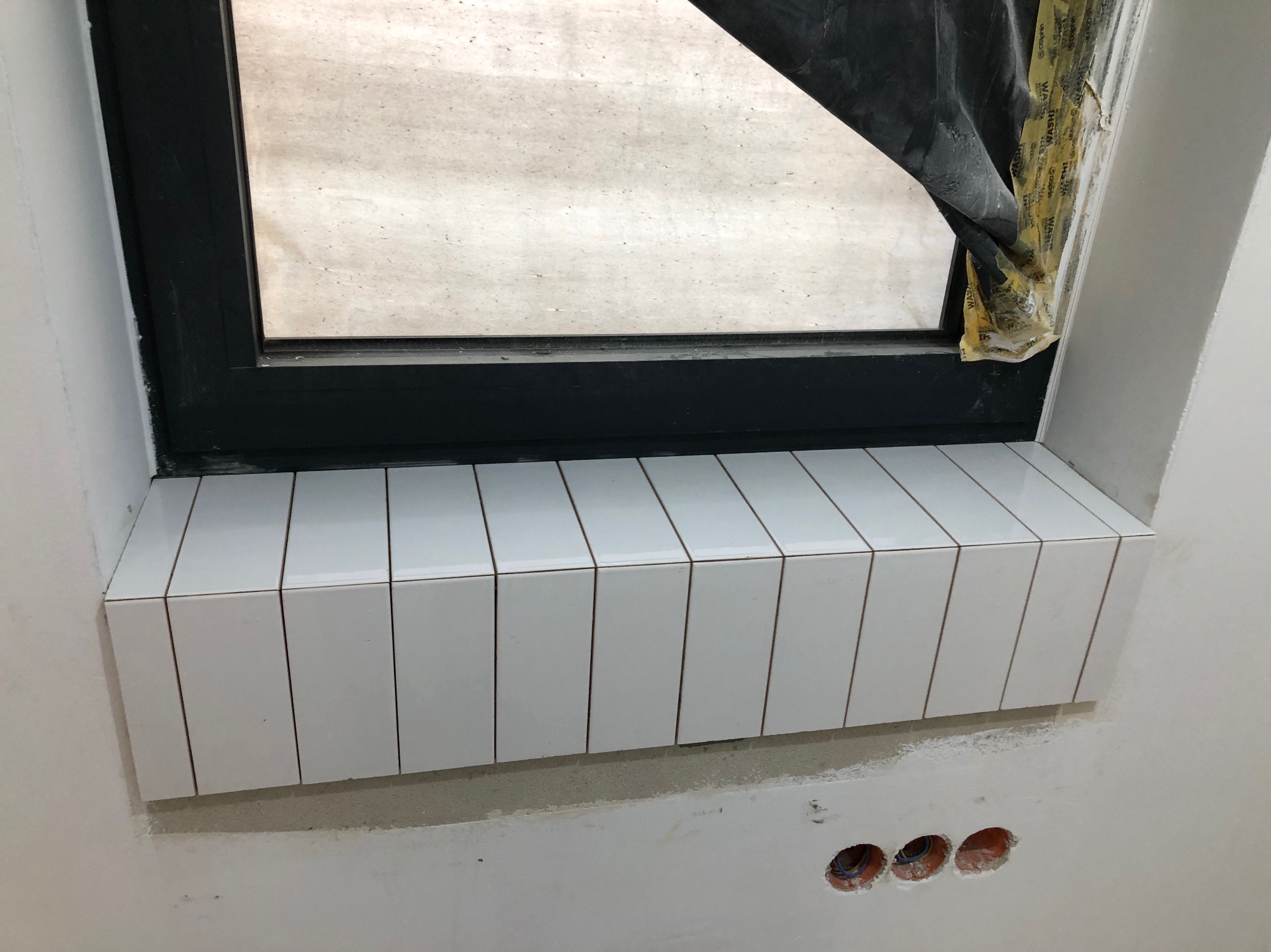 Białe, wąskie płytki ułożone na parapecie okna z czarną ramą. Fragment ściany z widocznymi otworami na gniazdka elektryczne. Okno zabezpieczone folią malarską.