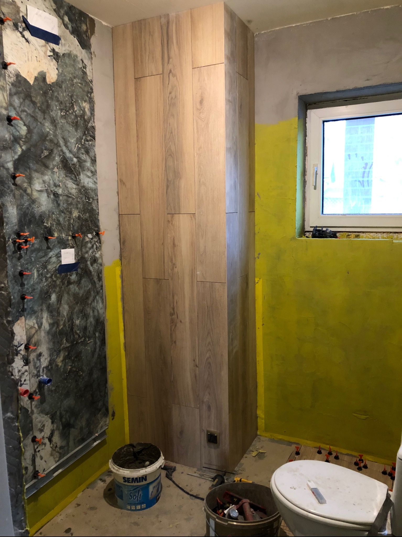 Remont łazienki w trakcie: obłożona drewnopodobnymi płytkami pionowa zabudowa, fragment ściany z szarą płytką i żółtą hydroizolacją, widoczne narzędzia i akcesoria glazurnicze.