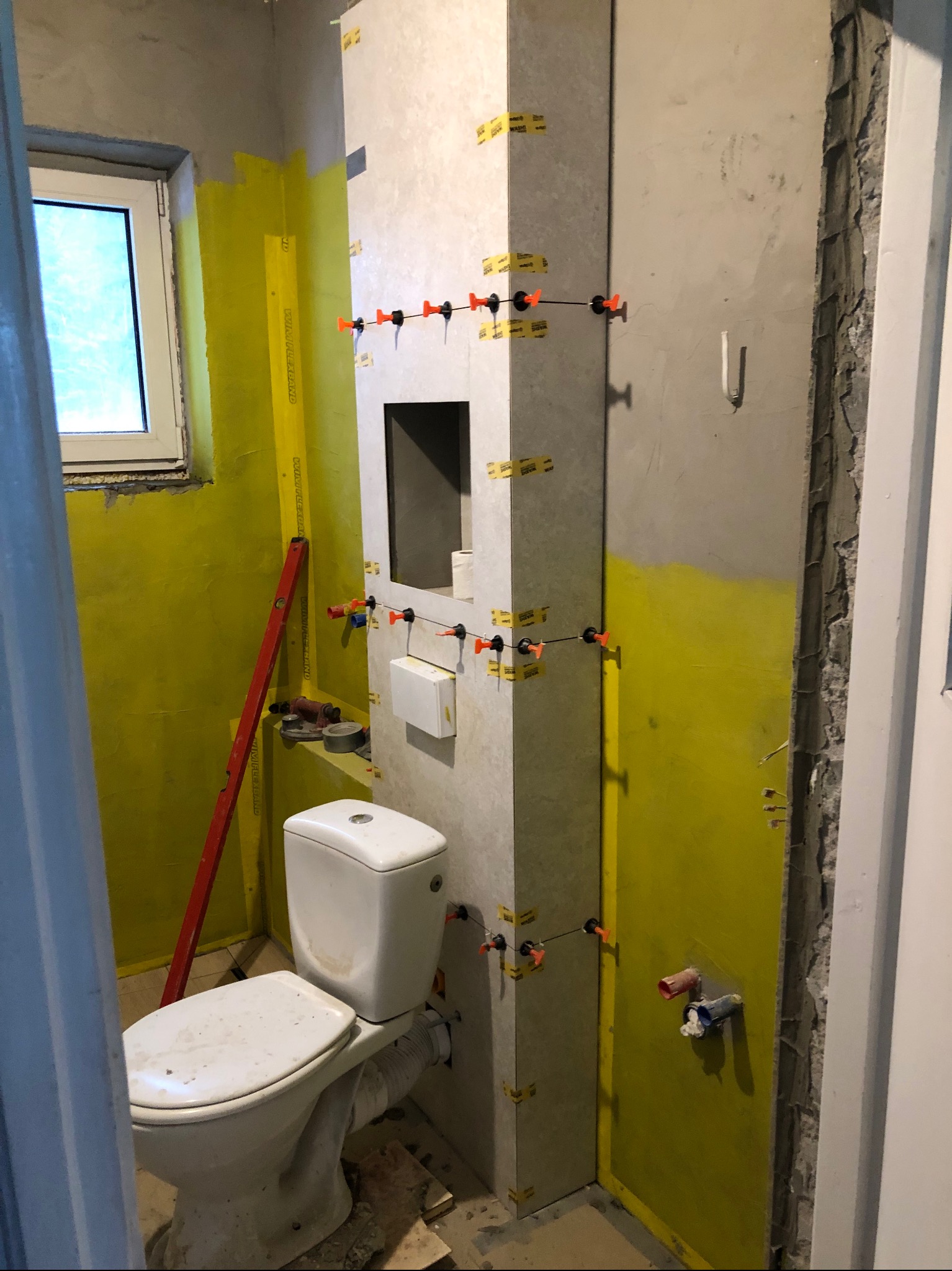 Łazienka w trakcie remontu: ściany z żółtą hydroizolacją, częściowo ułożone płytki z systemem poziomowania, widoczna toaleta i poziomica. Prace wykończeniowe w toku.