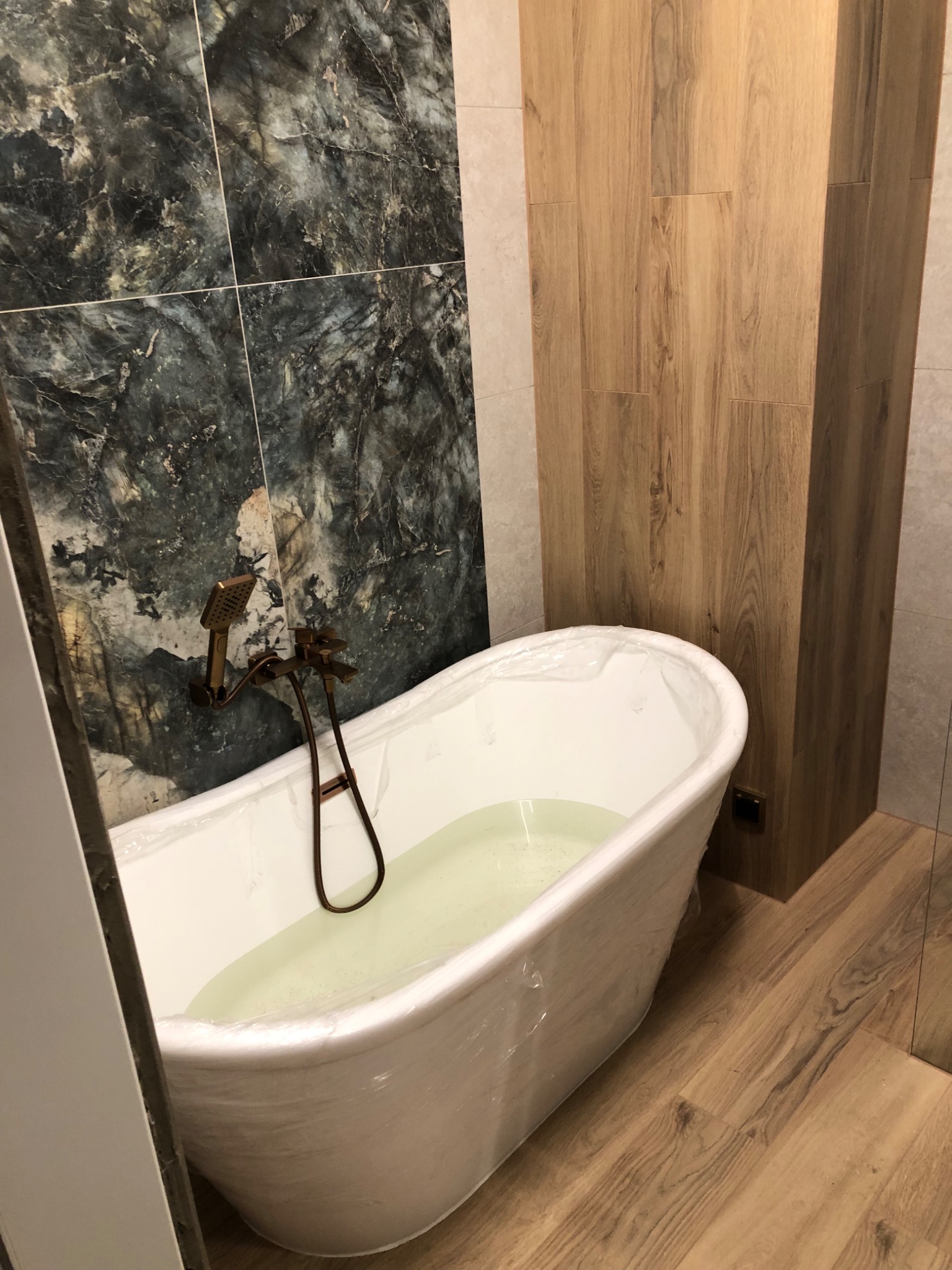 Wolnostojąca wanna w folii ochronnej, bateria w stylu retro, ściana z płytek imitujących marmur i drewnopodobna okładzina. Nowoczesna aranżacja łazienki.