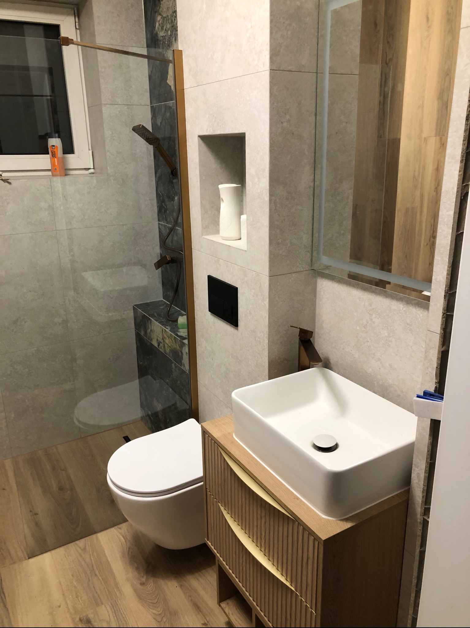 Nowoczesna łazienka po remoncie: kabina prysznicowa ze złotymi akcentami, umywalka nablatowa na szafce z lamelami, toaleta podwieszana i płytki imitujące drewno na podłodze.