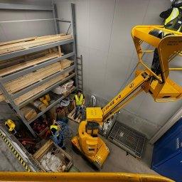 ANMACO KAMIL MAK - Praca na wysokości w magazynie: żółty podnośnik koszowy z pracownikami w kamizelkach odblaskowych, instalacja kabli przy regałach z drewnem. Widok z góry.