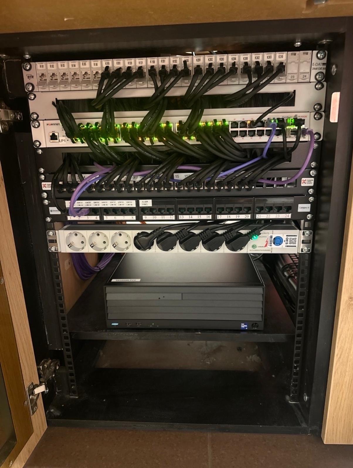 Szafa rack z patch panelem, switchami i listwą zasilającą, uporządkowane okablowanie sieciowe wewnątrz drewnianej obudowy, widoczne porty RJ45 i kable Ethernet.