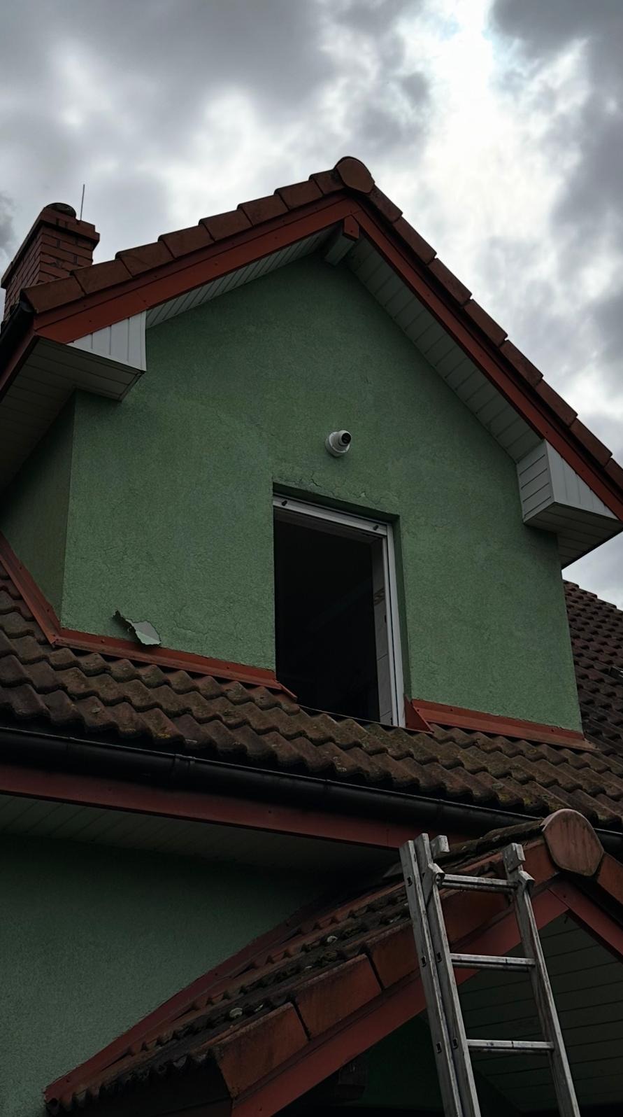 Zielony dom z brązowym dachem i kamerą na ścianie, widoczna drabina. Ujęcie z dołu, pochmurne niebo w tle. Otwór okienny bez okna.
