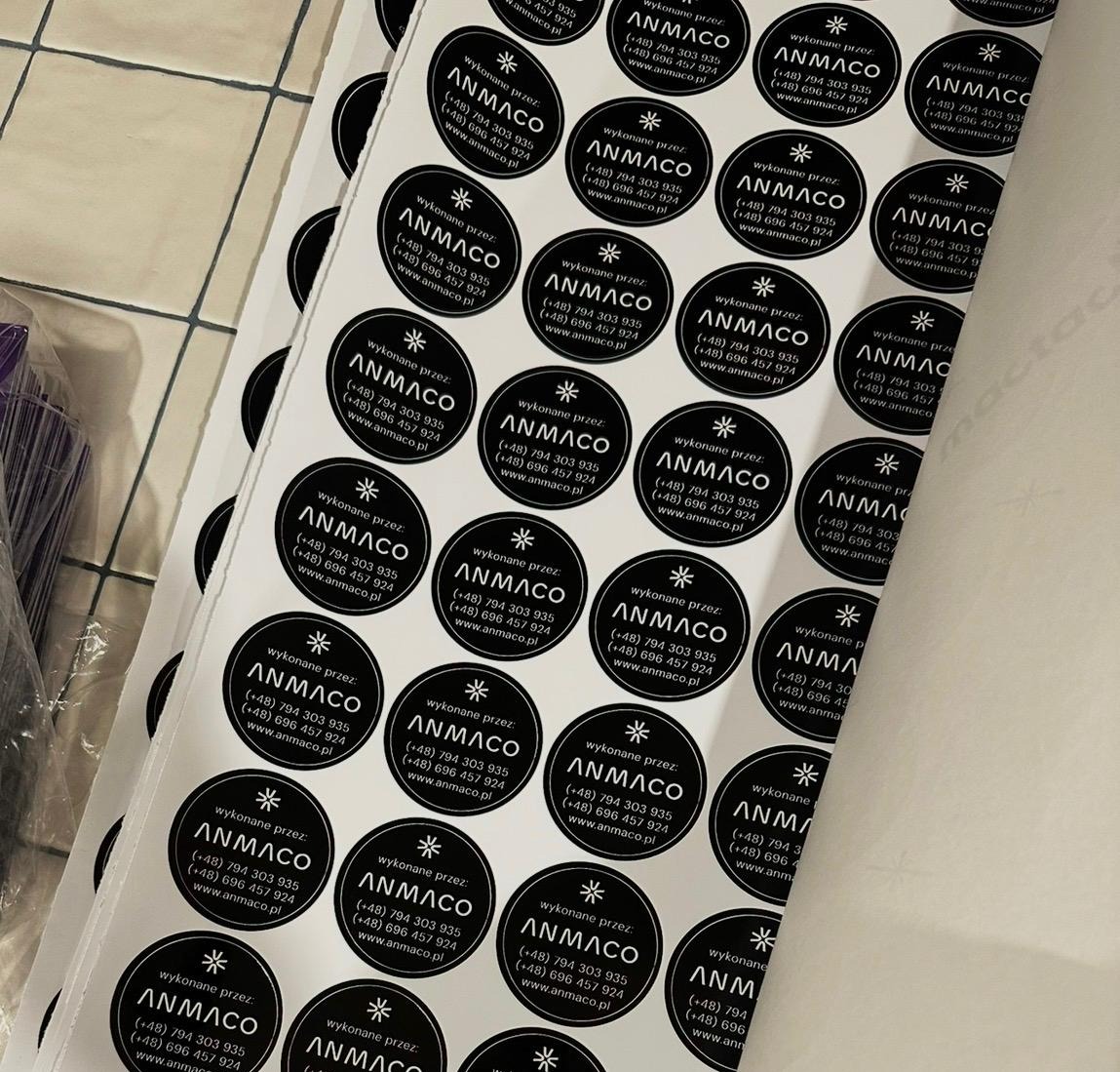 Arkusz z okrągłymi naklejkami z logo i danymi kontaktowymi firmy ANMACO, widoczne numery telefonów i adres strony internetowej. Naklejki w czarno-białej kolorystyce.