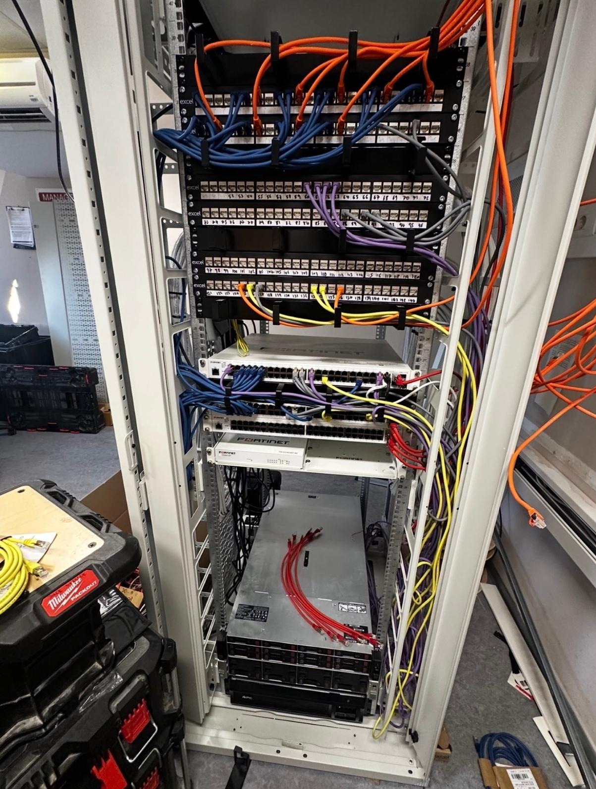 Szafa rack z patch panelem, switchem i serwerem Fortinet. Kable sieciowe w kolorach: pomarańczowy, niebieski, żółty i fioletowy. Widoczne narzędzia Milwaukee.