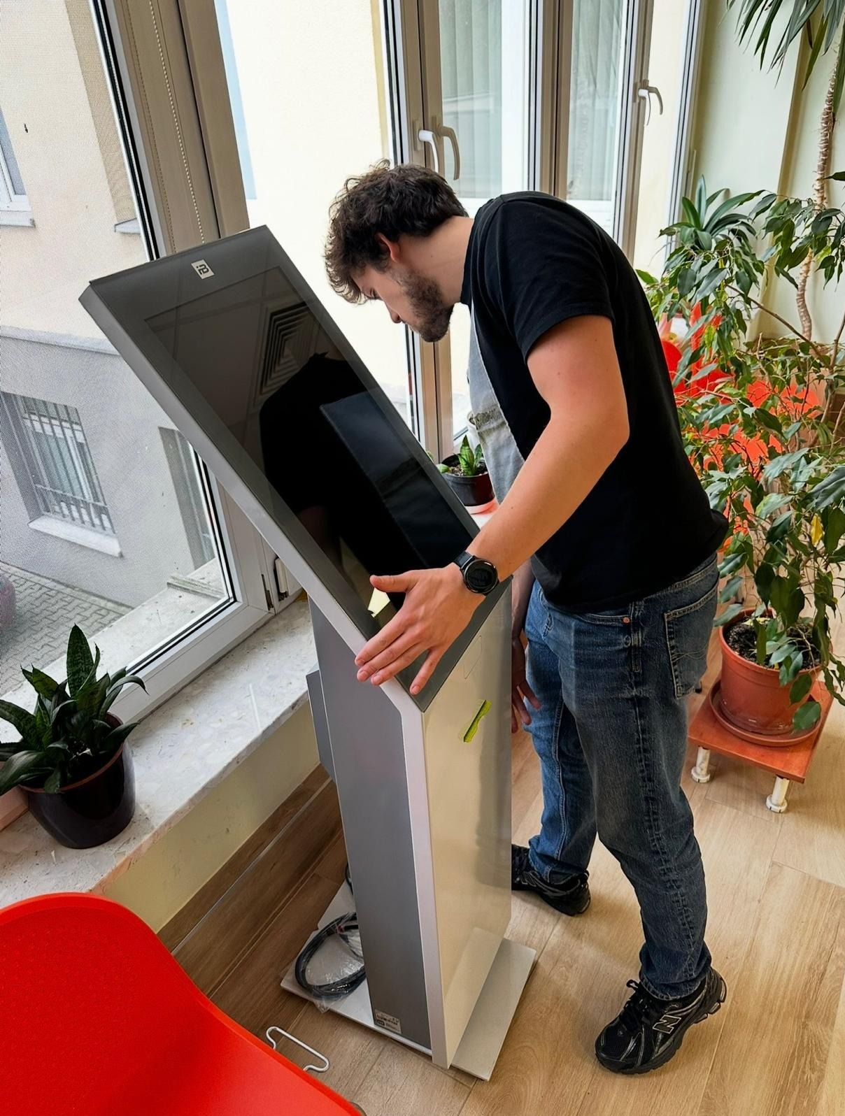 Mężczyzna testuje interaktywny kiosk informacyjny z ekranem dotykowym w jasnym pomieszczeniu z roślinami doniczkowymi i oknem w tle.